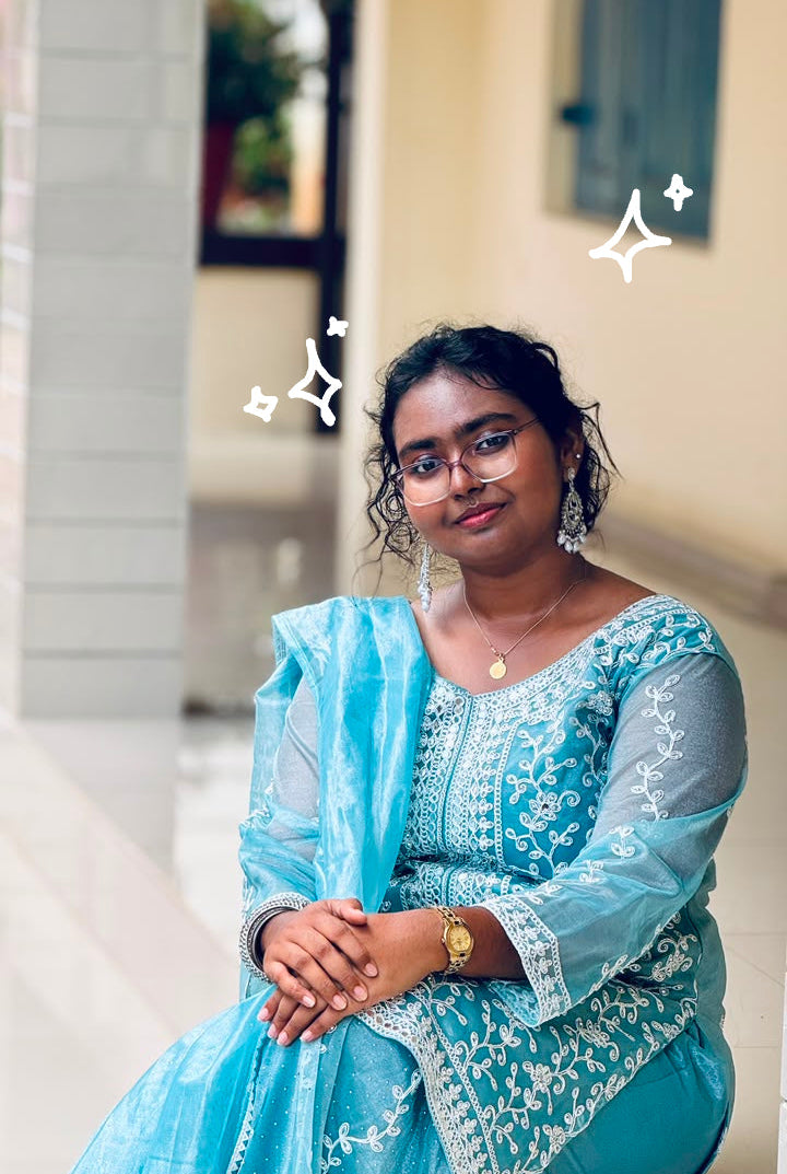A woman wearing a light blue traditional embroidered outfit with sheer sleeves and a matching dupatta, seated in an indoor corridor with beige walls and columns.