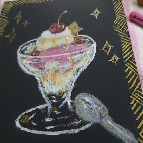A colorful chalk-style drawing of a dessert sundae on a black background with gold decorative borders, surrounded by pastel crayons and a paintbrush on a pink striped fabric surface.