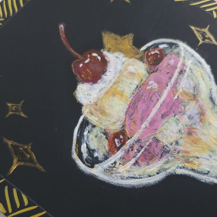 A close-up of a black square art print or card featuring a hand-painted ice cream sundae in a glass with whipped cream, cherries, and gold decorative star and stripe patterns on a pink and beige background.