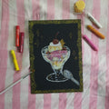 A square black art print or card featuring a hand-drawn ice cream sundae in a glass with a cherry on top, surrounded by crayons and a paintbrush on a pink and white striped fabric background.